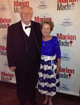 The couple smiles in front of a MarionMade! backdrop. Chuck wears a black tuxedo with a black bowtie. Pam is shorter with a blue top and white and blue silky skirt to her ankles.