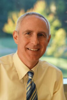 A white man with short white and gray hair, a big smile, a yellow shirt and a blue striped tie.