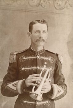 A man wears a band uniform and poses while holding a coronet. 