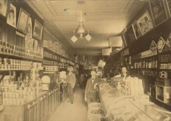 A black-and-white photo of the inside of the store, featuring barrels of fruit, canned goods, and cases. 