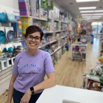 Lindsey Henry smiles while leaning on the counter in her toy store. She has short black hair, dark glasses, and a smile.