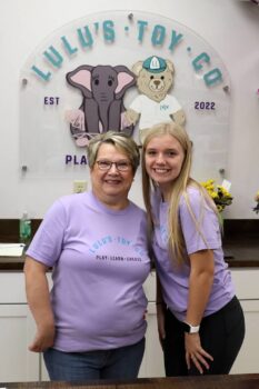 Two employees smile in front of the logo. 