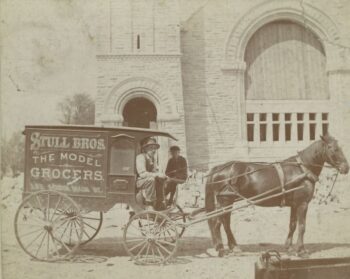 A man drives a horse-drawn wagon with the name "Stull bros the model Grocers"