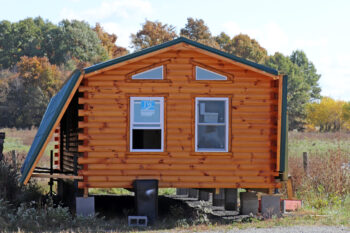A log cabin with reddish wood logs and four windows sits on blocks. the roof folds down and the porch folds up to transport it on semis to its final location.