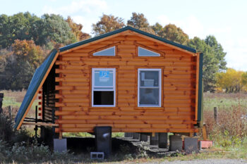 A log cabin with reddish wood logs and four windows sits on blocks. the roof folds down and the porch folds up to transport it on semis to its final location. 