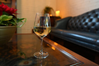 A glass of a white wine stis on a coffee table. A black leather couch is in the background along with white brick walls and candles. 