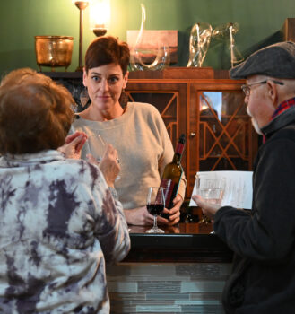 Megan Dawson, a tall white woman with short brown hair and a light gray sweater, holds a wine bottle behind the counter while she talks to two customers. 