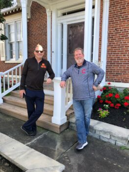The two brothers pose in front of the True home and smile. Jm is taller with short brown hair, snglasses, a Cleveland Browns zipped pullover and dark blue jeans. Rex has light brown hair, a gray beard, a gray Cincinnati Reds zipped pullover and light blue jeans. The brick home with a white porch and steps is in the background.