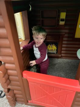 A toddler with short bonde hair, a red and white shirt and red pants plasy in a toy log cabin with a red swinging door.