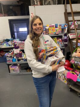 Volunteer and local teacher jane Harper, a white woman with long brown hair, a gray sweatshirt, and light blue jeans, holds an armload of puzzles as she helps someone shop for a child in need.