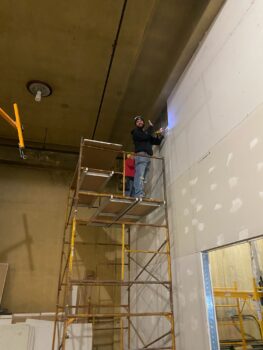 Two men on a scaffolding work to install a sheet of drywall.