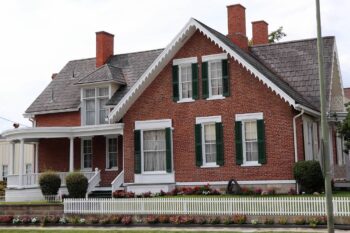 The two-story home has red brick, blac shutters, and white trim. There is a white picket fence out front. 