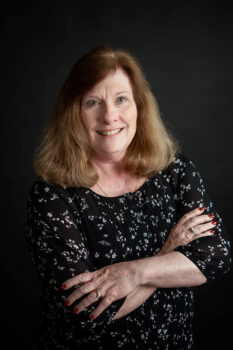 Jean smiles at the camera. She is a white woman with shoulder length light brown hair. She has one blue eye, one green eye, a smile and a black and white patterned shirt. She is crossing her arms. 