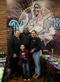 The family smiles in front of a giant Elvis Pigsley mural (a pig in a white jumpsuit with Black Hair, glasses, and a Microphone. Shannah is a white woman with a bony tail. Robert is a taller black man with a gray and black hair and beard. Sierra is a young Black woman with a wide smile and pulled back hair. Martin is a handsome elementary school-aged boy with short black hair, brown skin, a smile, a Spiderman t0-shirt, red and black flannel shirt, jeans and tennis shoes.