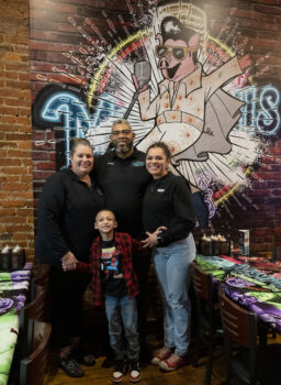 The family smiles in front of a giant Elvis Pigsley mural (a pig in a white jumpsuit with Black Hair, glasses, and a Microphone. Shannah is a white woman with a bony tail. Robert is a taller black man with a gray and black hair and beard. Sierra is a young Black woman with a wide smile and pulled back hair. Martin is a handsome elementary school-aged boy with short black hair, brown skin, a smile, a Spiderman t0-shirt, red and black flannel shirt, jeans and tennis shoes. 