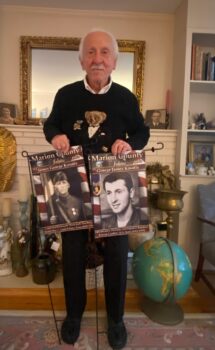 George smiles and holds the flags honoring his father and himself. Both of the flags display their photos in their uniforms and their regiments. 