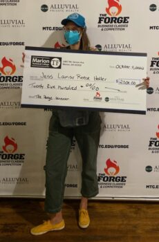 The winner holds a giant check for $2,500.She is a white woman with long brown hair, a blue hat, a blue mask, glasses, olive pants and yellow shoes.