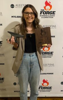 Allison Longsdorf holds her plaque and small anvil awards. She is a white woman with wavy, long brown hair, glasses, a bright smile, a black shirt, acid washed jeans and a tan suit coat.