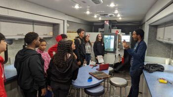 A college student speaks to a group of high school students. 