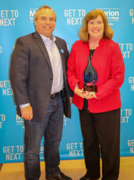 Kit Fogle Smiles while standing next to Jean Obenour as she holds her blue glass award. They stand in front of a blue backdrop with the Marion Technical College logo and the phrase Get to next in white lettering. 