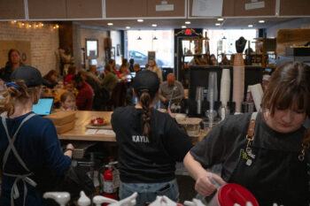 Three baristas work behind the counter at the small coffee shop in a busy rush. The coffee shop has a wooden counter, brick walls, large windows facing the street, and tables with customers.