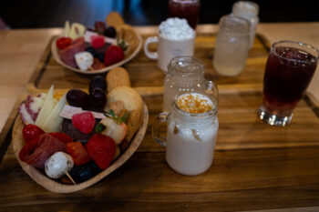 A wooden board hosts four different drinks in various Mason jars and glasses enxt to a bowl with meats, cheeses, fruits, crackers and olives. 