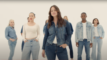 Karah smiles while wearing a faded denim jacket and white shirt with jeans. She has brown, long, wavy hair and a light pink lipstick in a slight smile. Other models wearing denim are in the background. 