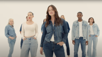 Karah smiles while wearing a faded denim jacket and white shirt with jeans. She has brown, long, wavy hair and a light pink lipstick in a slight smile. Other models wearing denim are in the background.