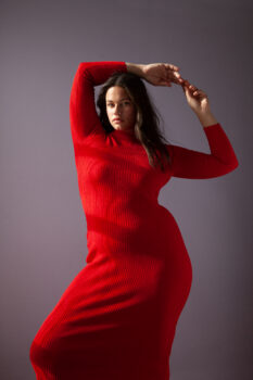 Karah looks at the camera while wearing a long red ribbed sweater dress She has long brown wavy hair, brown eyes, full lips and a cleft chin.