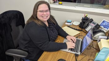 Katie sits at her desk. She is white woman with glasses, a wide smile, long, straight brown hair, a black and white polka dot shirt and a blck sweater.