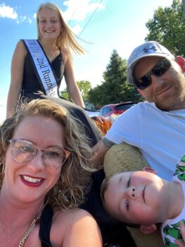 The family smiles while riding in a convertible in the Popcorn Parade. Kaitlin is a white woman with blonde, curly highlighted hair; bronze glasses, a silver stud piercing above her upper lip, and red lipstick, gold hoop earrings and a gold rope necklace. Lexi is a white teenage girl with long, straight light blonde hair and a smile. She wears a black gown with spaghetti straps, a silver necklace and a white sash stating "2022 Miss Teen Popcorn Festival 2nd runner up." A young boy with blue eyes, sandy blonde short hair and rosy cheeks wears a Minecraft shirt. The father is a white man with a gray camo ballcap, black sunglasses, and a red beard. he has a white shirt and a sleeve of tattoos on his arm.