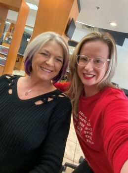 The client poses with Kaitlin after an appointment. Teena is a white woman has a chin-length blonde bob with dark purple tones at the bottom. She has a wide smile, a delicate gold necklace with a pendant, and a black sweater. Katilin has long, wavy blonde hair, clear-rimmed glasses, a piercing above her lips, red lipstick and a red shirt. The salon is visible behind them. 