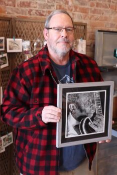 Brian holds one of his framed pieces of artwork inside the store. He is a white man with a short gray beard, glasses, and a red flannel shirt over a T-shirt.