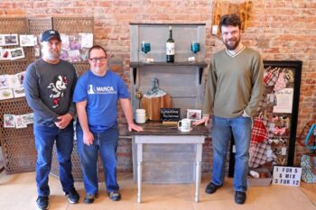 Decorative. Randy is a tall older white man with a ball cap and gray beard with a serious expression. Bobby is shorter, young, white man with short brown hair, black-rimmed glasses and a smile. Nick is a taller young, white man with curly brown hair and a brown beard and a bright smile. They pose next to a shelf made from an old door that has been painted gray. It has a higher shelf affixed to the front holding a wine bottle and a larger shelf at the bottom with decorative wooden legs in the front and the doorframe as the back legs. 