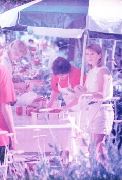 A young woman with dark blonde, shoulder-length straight hair, a white sleeveless shirt, khaki shorts and a apron sells strawberries at a farmer's market