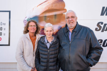 The family who owns and operates the G&R stands outside the building. Misty is a woman with brown curly hair, glasses, an orange shirt and a cream fleece jacket. Joy is a shorter, older woman with short white hair, a smile,m a gray shirt with a collar and a puffy black jacket. Bernie is a tall man with short white hair, glasses, a white beard, a blue button-down shirt and a black leather jacket.