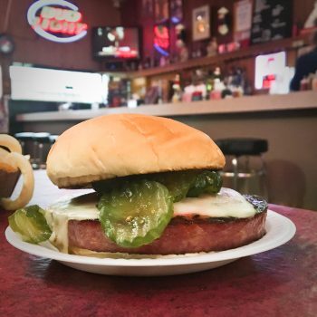 The famous bologna sandwich is a 1' thick slice of bologna with melted white Monterey Jack cheese, pickles, raw onion, and a soft hamburger bun. The bar is in the background.
