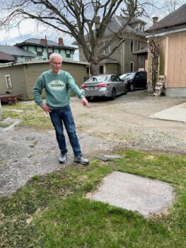 Ron stands behind the DeWolfe house and points to a large metal plate, perhaps 2.5' x 4', which covers the cistern. Ron is a tall white man with hort white hair, black glasses, a green sweatshirt, jeans and tennis shoes.