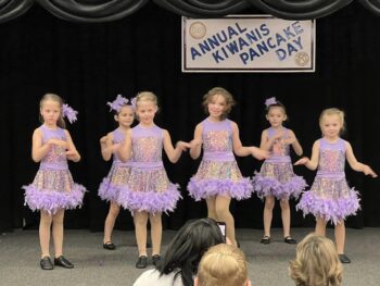 girls in shimmery lavender dresses addorned with purple feathers dance in unison on a stage at the event.
