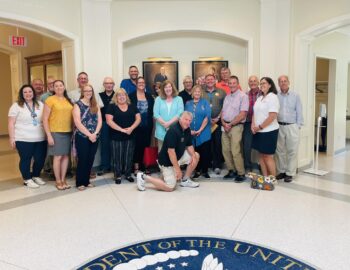 A group of about 20 Kiwanis members and guests smile. The presidential seal is visible on the floor. 