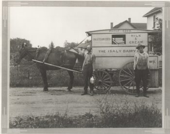 The black-and-white photo shows two men standing next to a wagon with "the Isaly Dairy Co. - Pasteurized milk & Cream; Phone - 4289" on the side. It is bulled by a black horse. The two white men wear long-sleeved, collared, button up shirts and dark pants. One man holds a wire basket with two milk bottles in it. One man has a hat and the other has a mustache. the dirt road is surrounded by wildflowers. Several houses are visible in the background.  