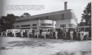 Rows of white and black motorized delivery vehicles stand outside the plant. The drivers standing next to each one have white short-sleeved shirts, white pants rolled at the cuff, and some have a black satchel or are holding a hat. The trucks and the building have "Isaly" on the sides. The factory is made of light bricks with rounded corners and block glass widows. 