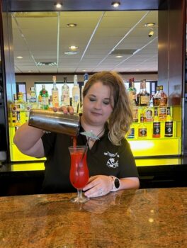 A white woman with blonde hair in a pony tail uses a silver shaker to pour a red drink into a curved glass with an umbrella.
