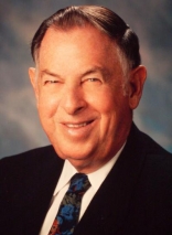 A photo of an older white man with a smile, short brown hair with white at the temples, and a black suit, white shirt and dark tie.