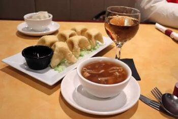 A bowl of hot and sour soup is placed next to a glass of plum wine next to an appetizer of crab rangoon. 