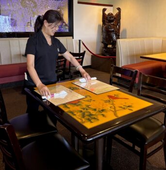 A server with long black hair in a pony tail smiles while putting silverware and menus on the tablle. A large glass feature with floating colored bubbles and other booths and tables, as well as a large Buddha statue, are in the background.