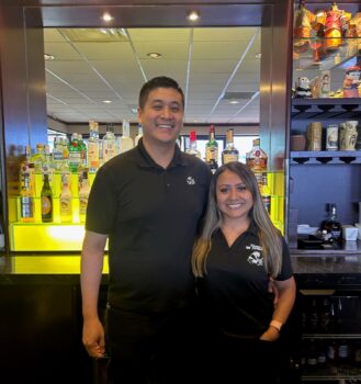 The couple stands in front of the bar. Frank is a tall man with short black hair and a smile. Jeny is shorter with long light brown hair . Both wear smiles. Their arms are around each other. 