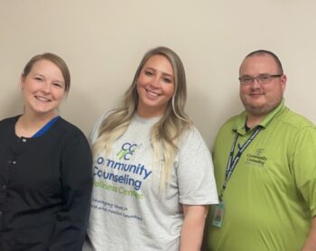The Crawford team members smile. Amanda is a white woman with dark blonde hair in a pony tail and navy scrubs covered by a black jacket. Lexi is a white woman long wavy blonde hair and a gray CCWC T-shirt. Dustin is a white man with buzzed, dark hair, facial hair and glasses. He is wearing a green CCWC polo and has a lanyard with his employee badge.