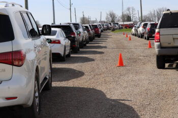 Lines of cars stand next to orange cones to receive free produce outside All Occasions.