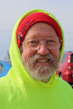 Jim is an older white man with a red knit hat, neon yellow hoodie, glasses, a gray beard and a smile.